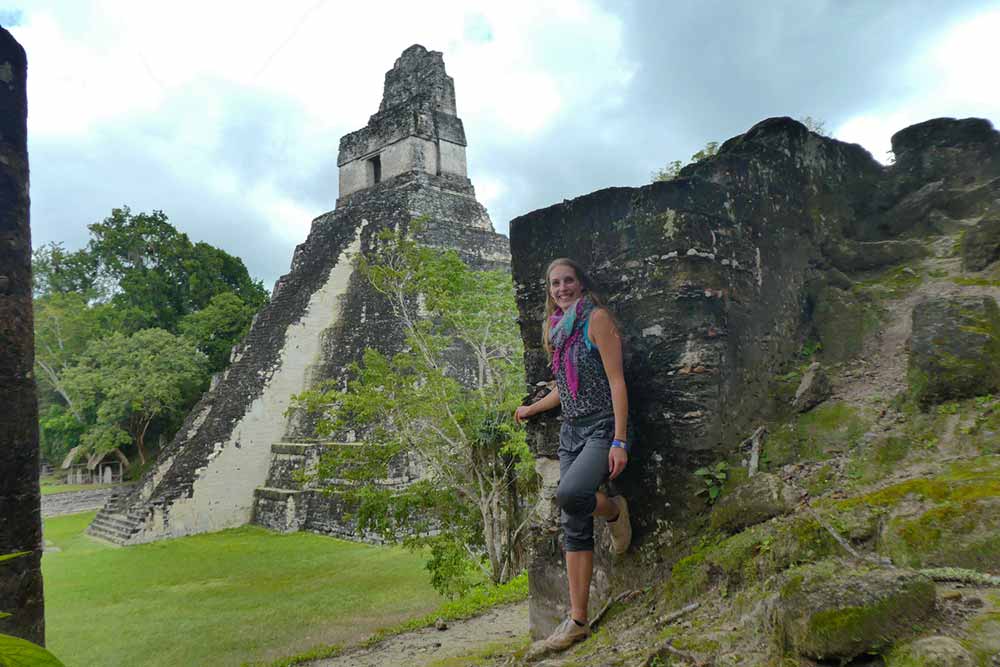A woman in a scarf is leaning on the wall of one of the Mayan Ruins of Tikal, with a clear blue sky in the background.