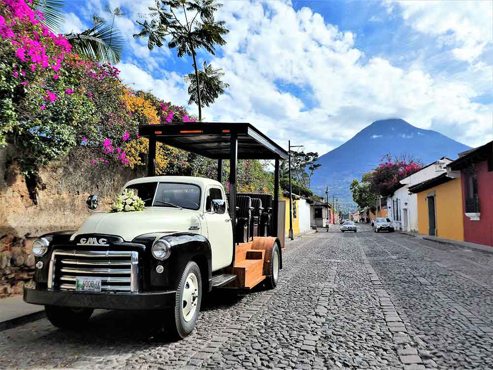 A white four-wheeled car is parked on the streets of Antigua, with a blue sky and mountains in the background.