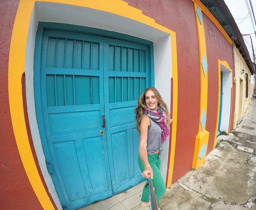 A woman with a scarf around her neck is taking a selfie amidst the vibrant and colorful houses of Flores