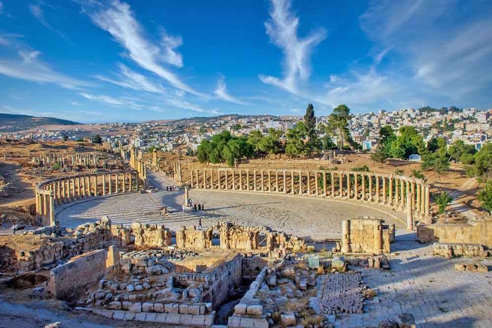 Oval Forum at Jerash features an open space in oval-shaped with a rows of towering columns at the side with cities and blue sky in the background