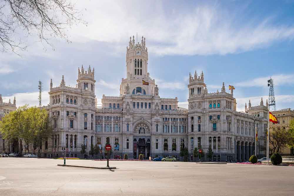 Plaza de Cibeles features a gray-white building with clock, multiple windows and intricate carvings stands against a blue sky