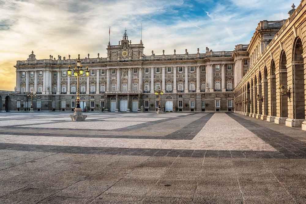 The Royal Palace of Madrid features a gray stone building with multiple columns and windows, standing against a blue sky.