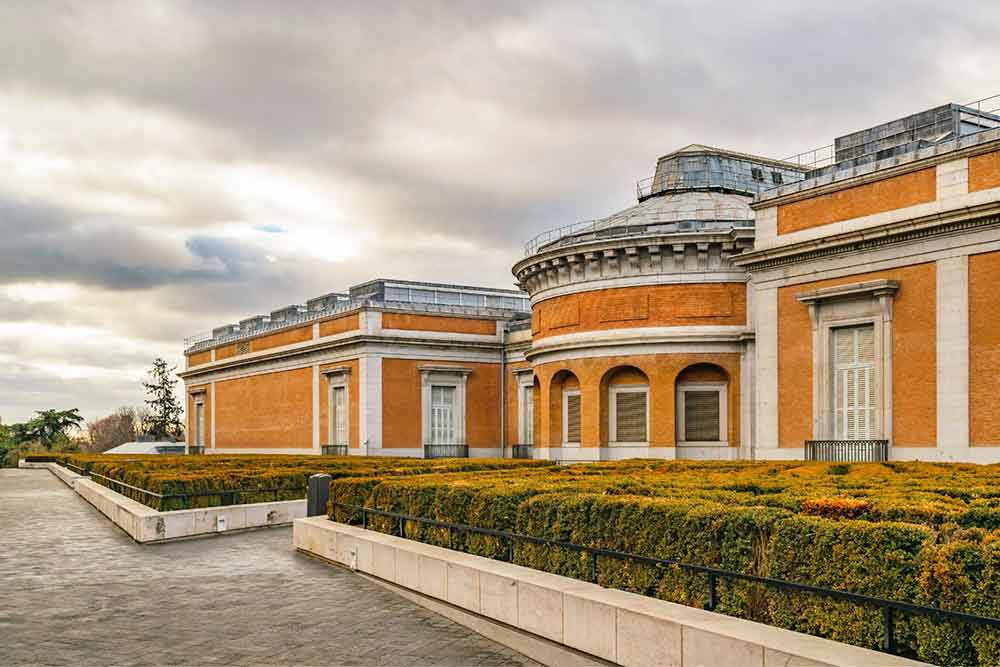 Prado Museum features a building painted in a red-orange color surrounded by plants 