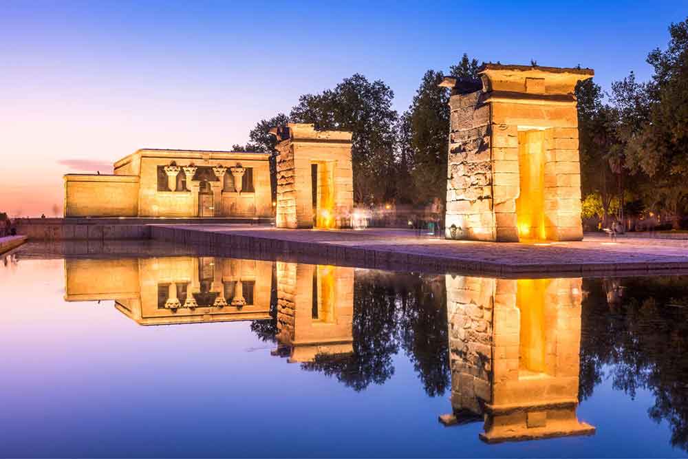 The Temple of Debod features a stone block standing in a row with yellow lights surrounded by trees and water
