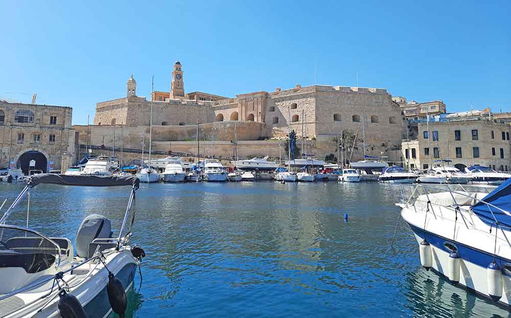 Plenty of white boats parked at the shore with a sandstone structure at the background in Cospicua Marina