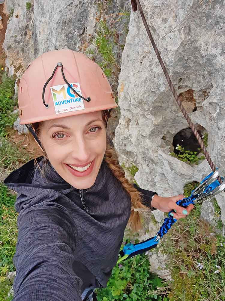 A woman wearing a red helmet is taking a selfie while preparing for Via Ferrata in Malta, equipped with a harness