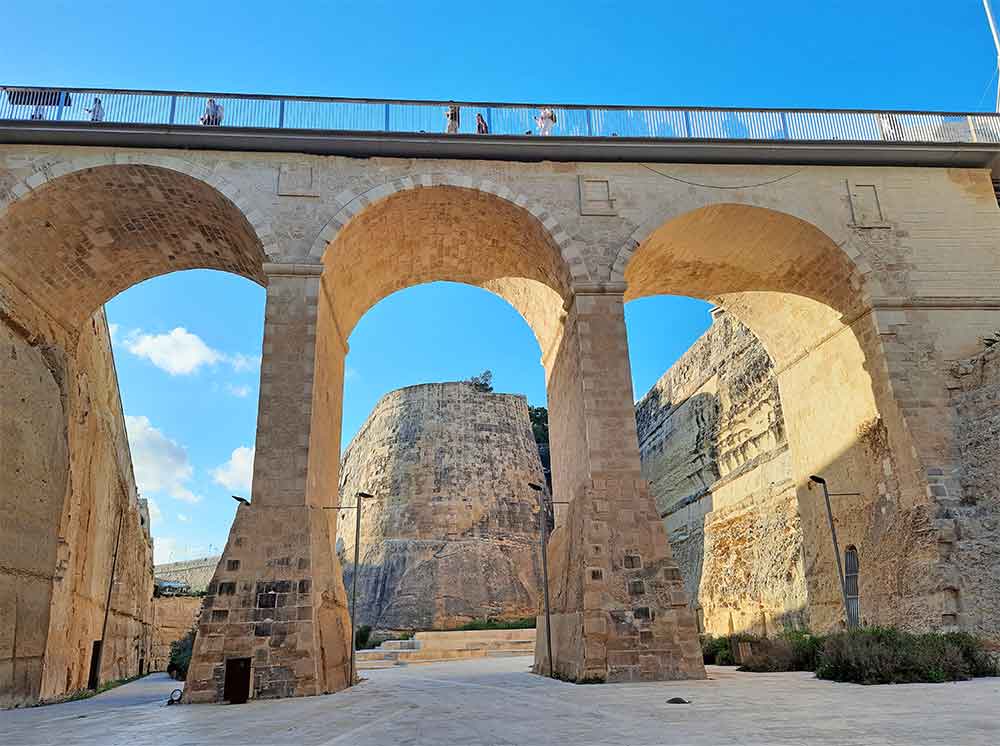 The city walls of Valletta showcase a tall brown bridge standing in the middle, with people walking by under a clear blue sky