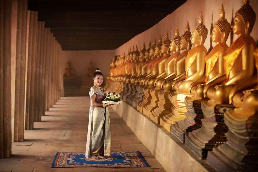 An Asian woman wearing a Thai dress is holding offerings inside the temple