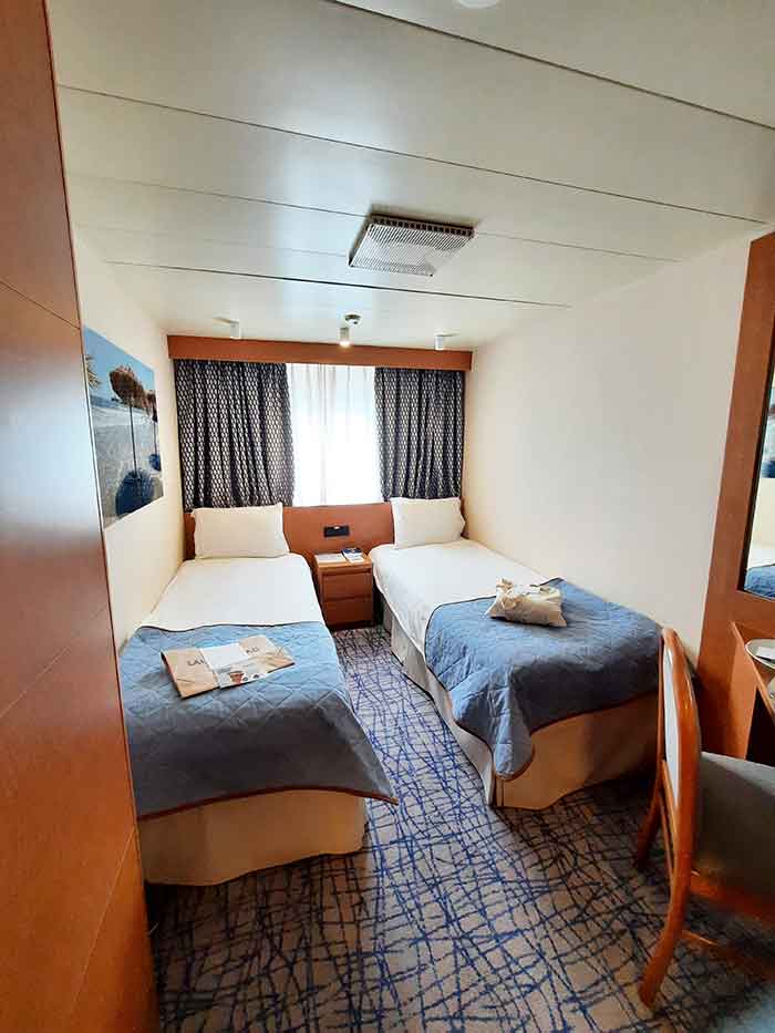 My cabin on my cruise. It shows two beds with white and clue covers and a window looking out to the sea at the back of the room. 