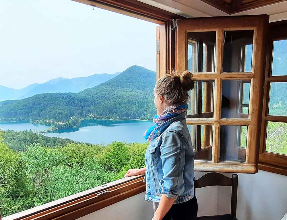 A woman is standing beside the window, looking at the view of Lake Doxa, surrounded by lush green trees and mountains