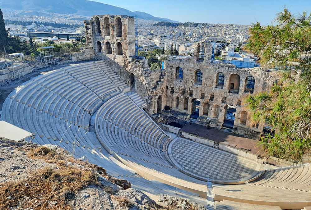 The Odeon of Herodes Atticus at the Acropolis is an open-air structure with a sandstone staircase and a front wall offering a stunning view of the city