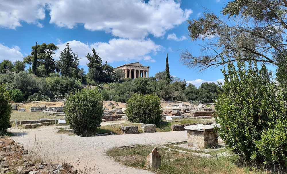 The Ancient Agora features columns and a roof in an earthy color, all surrounded by trees and plants.