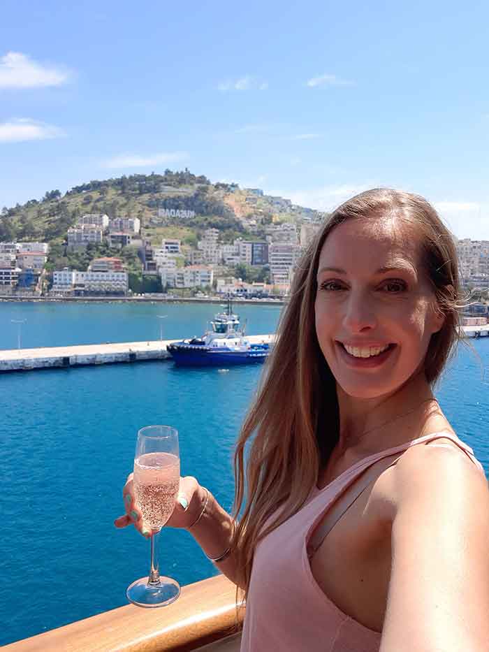 A woman (me) in a pink top holding a glass of pink champaigne. She is stood on the deck of a cruise ship. The cruise ships is in port and you can see other boars in the background and the town of Kusadasi in Turkey. 