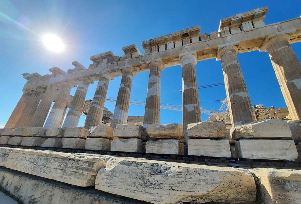 In the morning, the sun shines on The Parthenon, illuminating the multiple columns standing in a row and the large stone, all against a clear blue sky