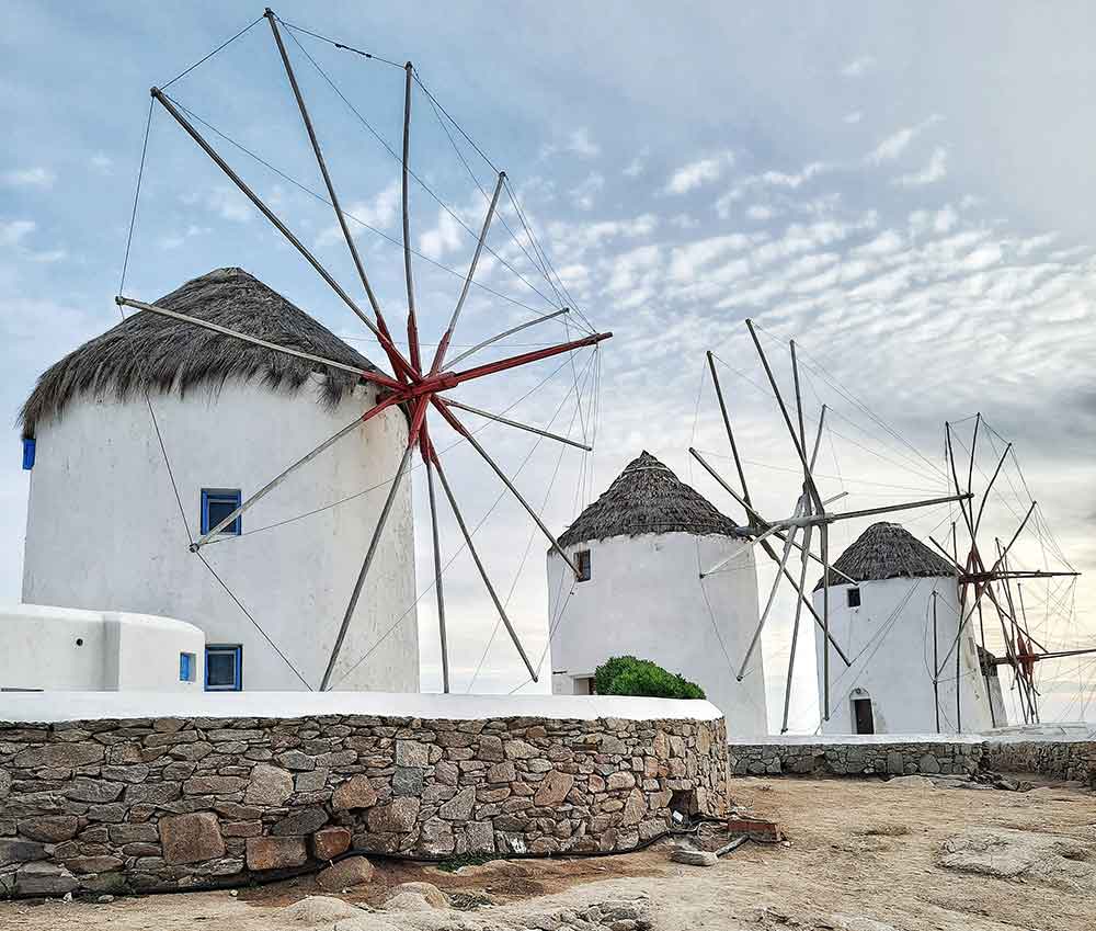 Three windmills are standing on Mykonos Island, each with a cone-shaped roof, all under a white-blue sky