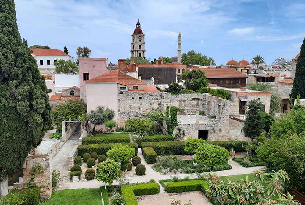 The view of Rhodes from the City Wall Walk features a manicured garden, plants, and trees surrounded by houses