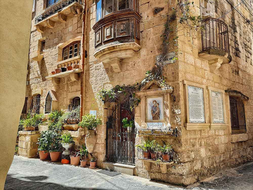 Streets of Rabat, Malta A sunny narrow lane in Rabat, Malta, showing a honey‑coloured stone building with a wooden enclosed balcony and wrought‑iron gate at the corner. Potted plants line the entrance, and a small religious shrine is nestled into the wall, all bathed in gentle winter sun in Europe.