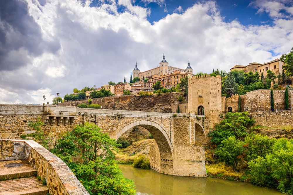 Discover how to visit the beautiful medieval city of Toledo. This easy day trip to Toledo from Madrid can be done by car, train, or organised tour. The image shows a view of the outside of the city with an old stone bridge going over a river.