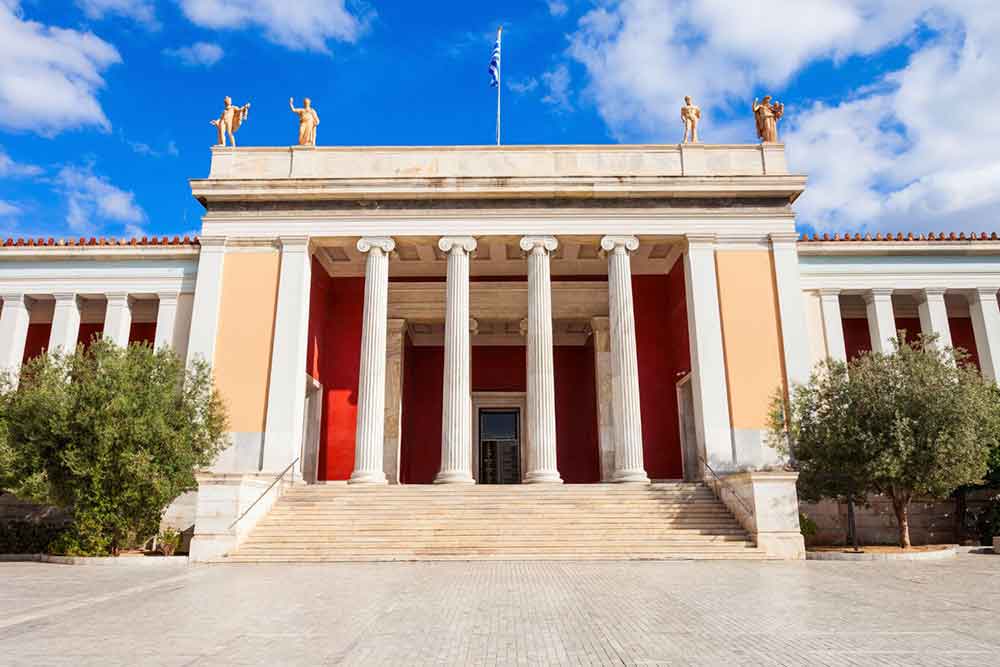 The National Archaeological Museum of Athens is a building with four columns and a red painted wall at the entrance along with 4 statues standing on the roof.