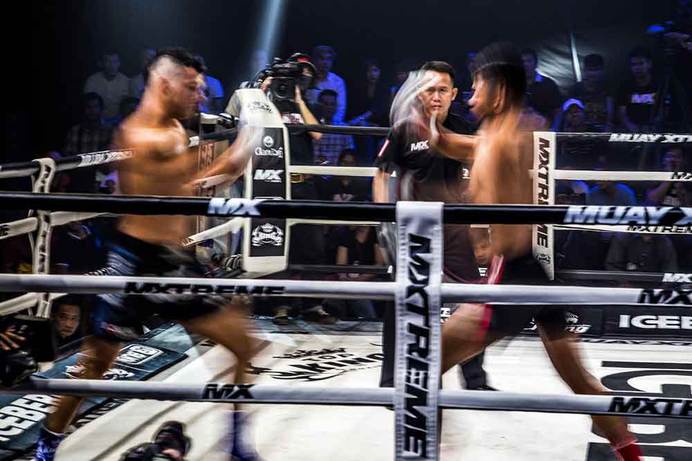 A live Muay Thai boxing match features two men wearing gloves engaged in a fierce fight inside the ring, with the referee overseeing the match, in front of a crowd watching the match