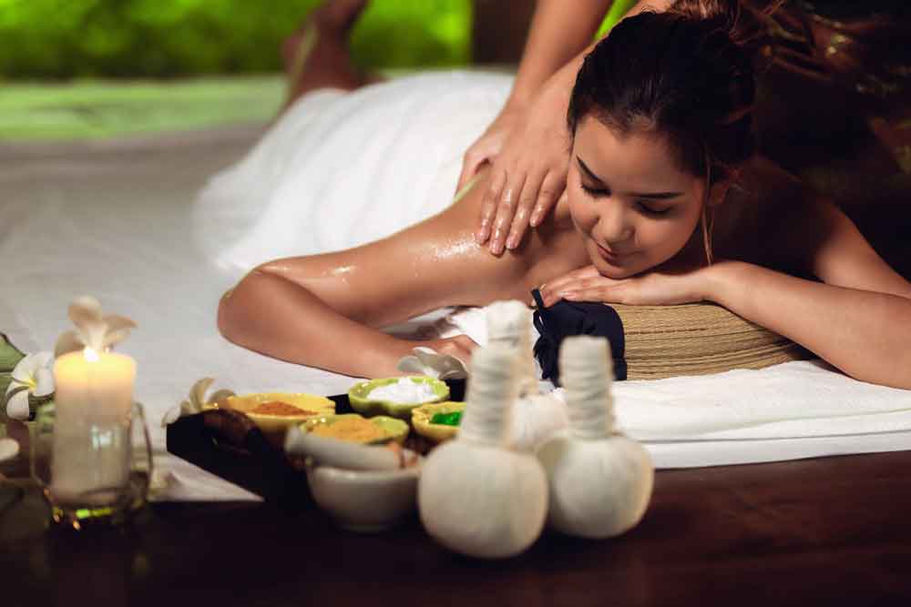 A woman lies on a mat on the floor, receiving a Thai massage with supplies and equipments at the front