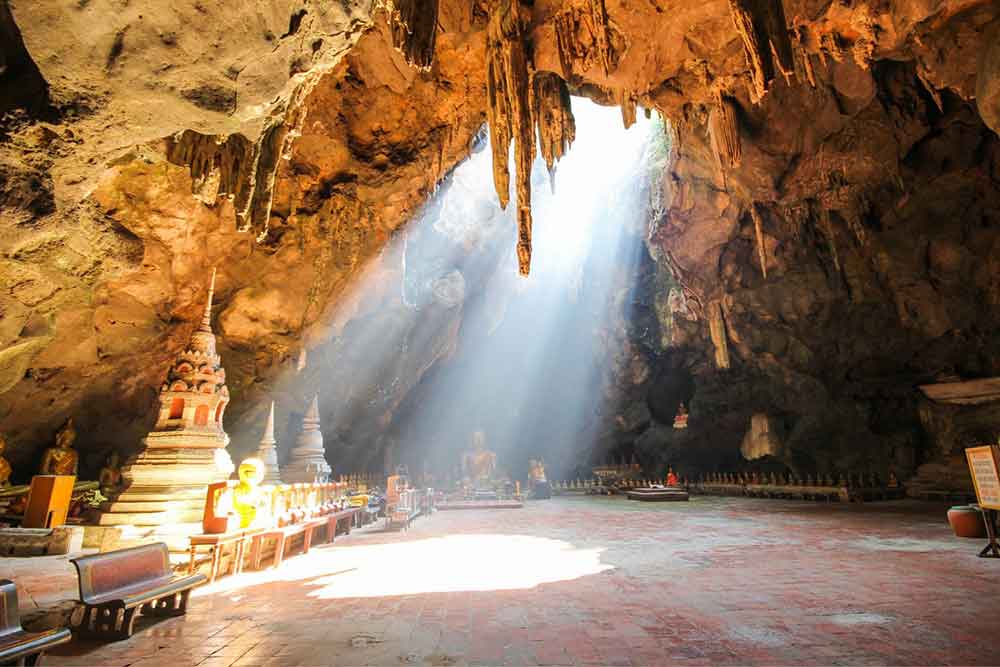 Khao Luang Cave at Phetchaburi, adorned with intricate carvings and illuminated by sunlight, showcases a collection of Buddha statues at its center