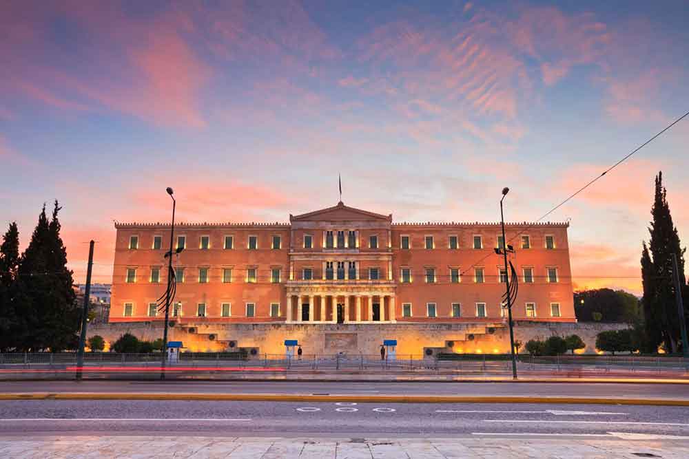 The Greek Parliament building is illuminated in Syntagma Square, with a sky painted in hues of blue and pink during sunset