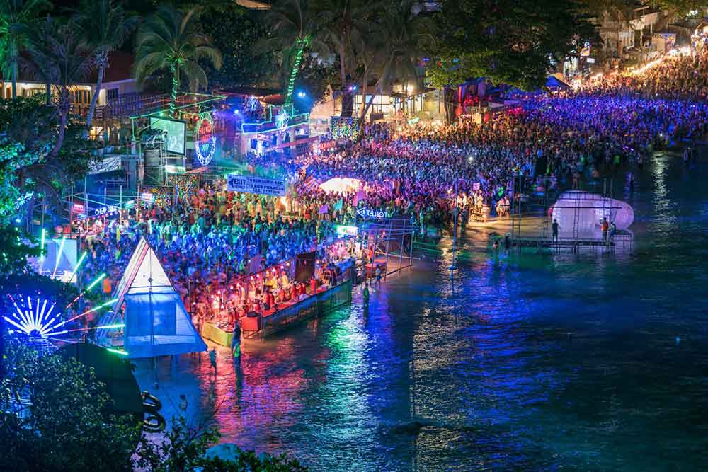 
A large crowd gathers at the Full Moon Party on Koh Phangan, with colorful neon lights and DJs creating a vibrant atmosphere at night alongside Haad Rin Beach