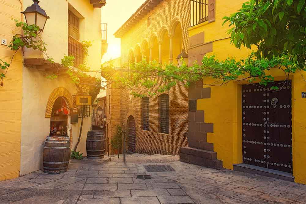 Barcelona Bucket List: 32 Best Things to Do in Barcelona, Spain 25 Street at Poble Espanyol, Barcelona, features yellow and brown houses, with sun rays shining down