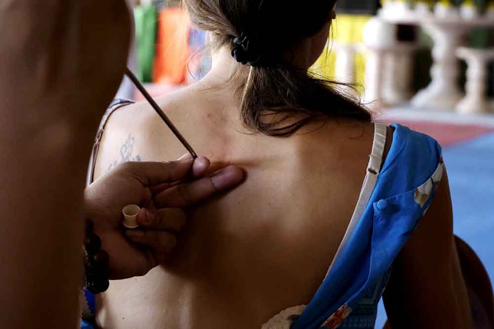 A woman is getting a Sak Yant tattoo using a bamboo stick at the back of her body