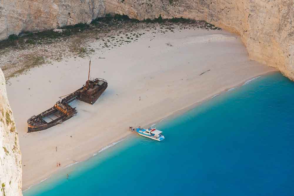 Emerging from the crystal blue water, the abandoned ship showcases a skeletal frame of rusted metal and broken panels, standing on the sandy shores of Zakynthos, surrounded by towering cliffs