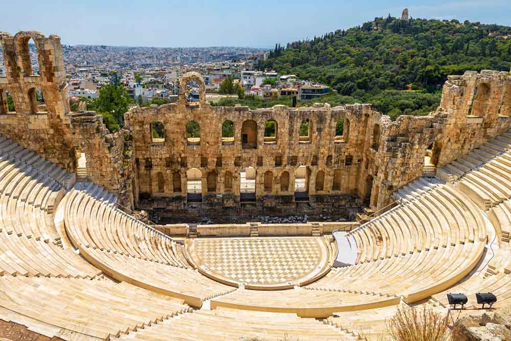 An open-air stone theater in a white-cream color seats, featuring a front wall that offers a majestic view of the city