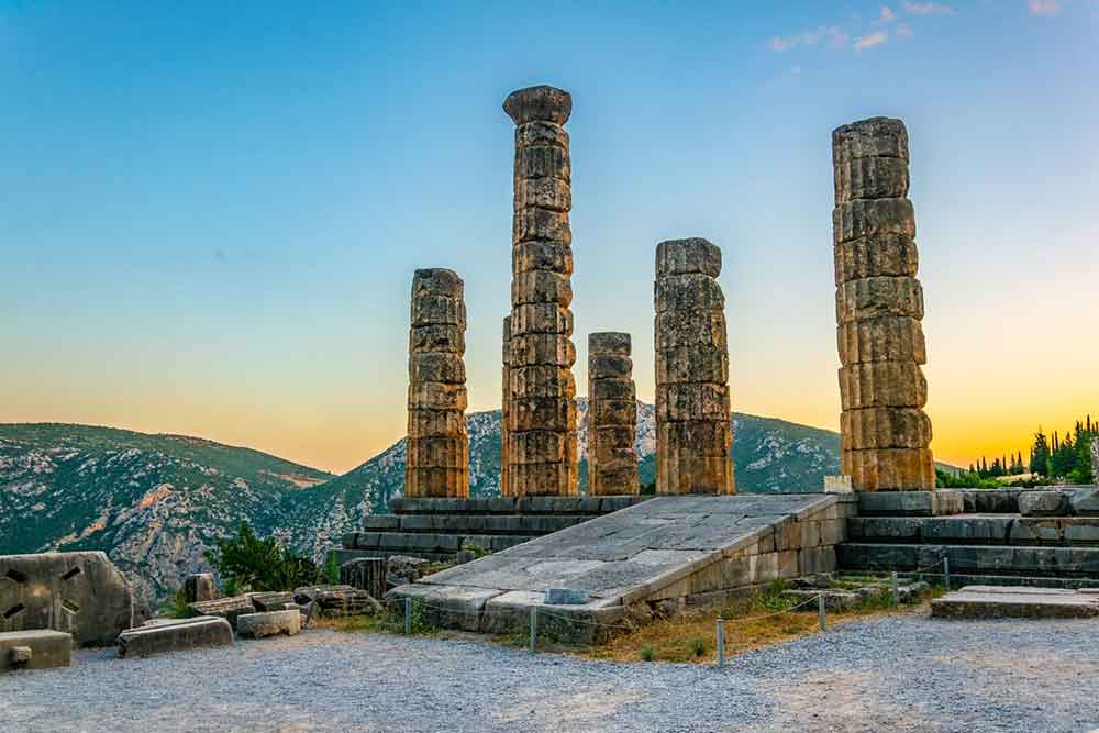 The ruins of the Temple of Apollo at Delphi, Greece, stand with sturdy columns made of limestone and marble in a black-white color, set against the serene backdrop of hills during sunset