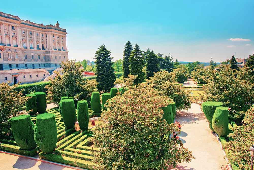 Sabatini garden in Madrid in a well manicured landsccape with lush green lawns, trimmed trees, and flower with the Royal Palace on the side