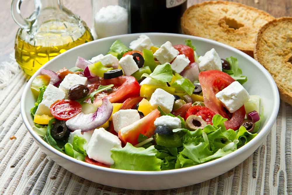 A bowl of Greek Salad with lettuce, tomatoes, onions, olives, feta cheese, oregano with olive oil in the side