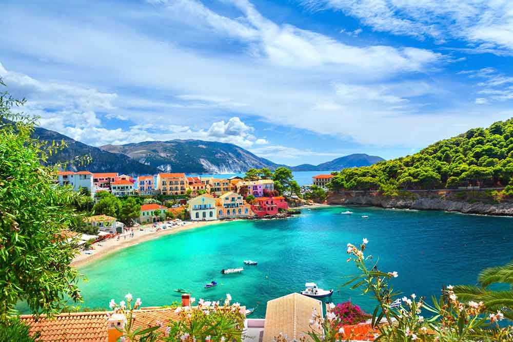 Village in Assos, Kefalonia, Greece features the beach in crystal-clear blue waters with boats, colorful houses, and mountains in the background, all under a clear blue sky 