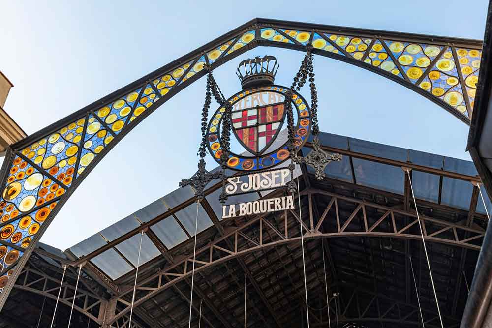 Barcelona Bucket List: 32 Best Things to Do in Barcelona, Spain 5 The arch at the entrance of Mercado de La Boqueria features a combination of yellow, blue, and orange under a clear blue sky