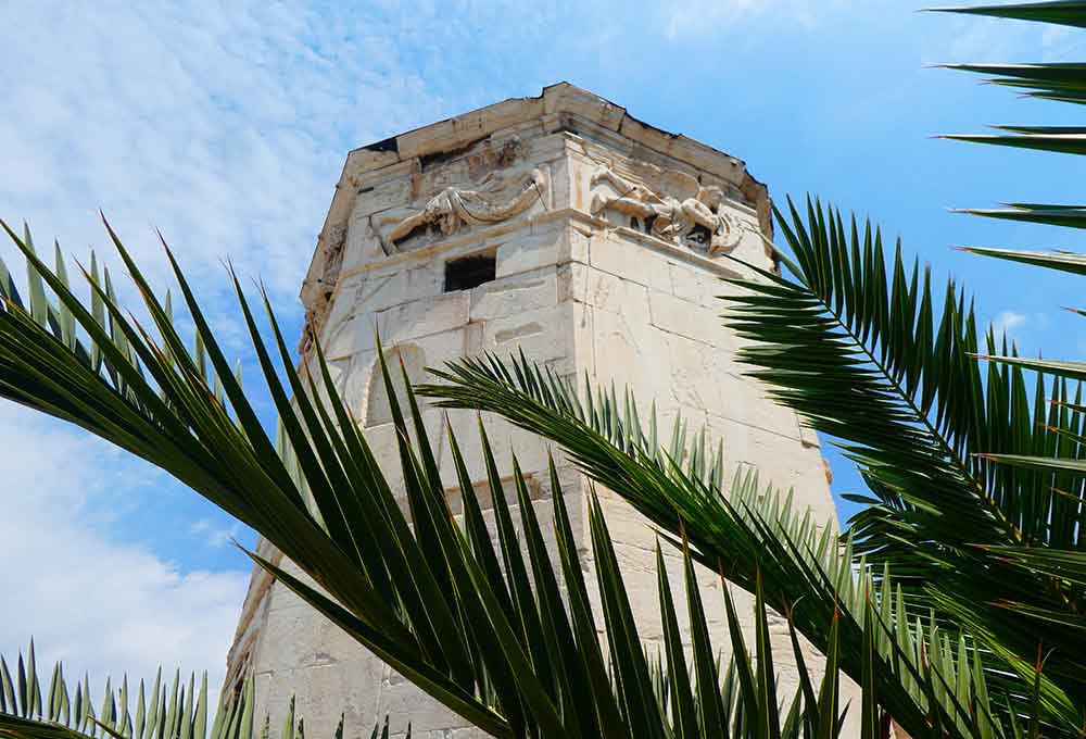 The Roman Agora features a magnificent marble tower with intricate designs and detailed sculptures, accompanied by a majestic palm tree in front with a clear blue sky