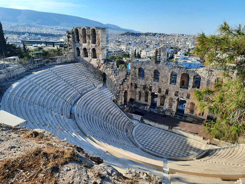 The Odeon of Herodes Atticus is an open-air stone theater structure, featuring a front wall that offers a panoramic view of the city and mountains against a clear blue sky