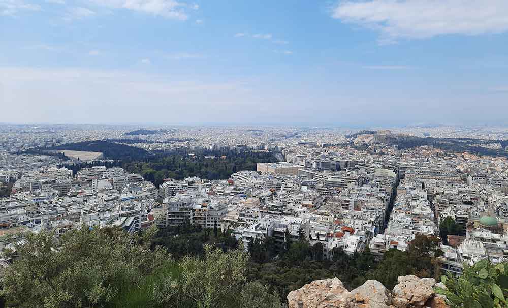 A panoramic view of the Athens cityscape showcases houses and trees seen from Mount Lycabettus during daytime