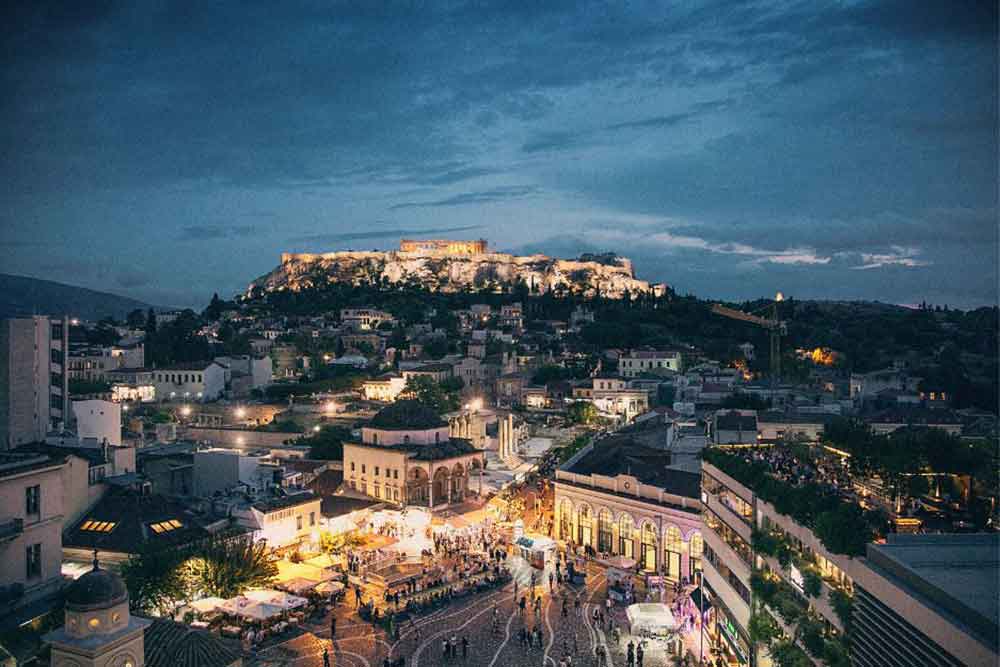 Acropolis at night offers a captivating sight with the lights emanating from nearby houses, creating a warm glow while on the ground, a group of people gathers, adding a lively atmosphere to Acropolis