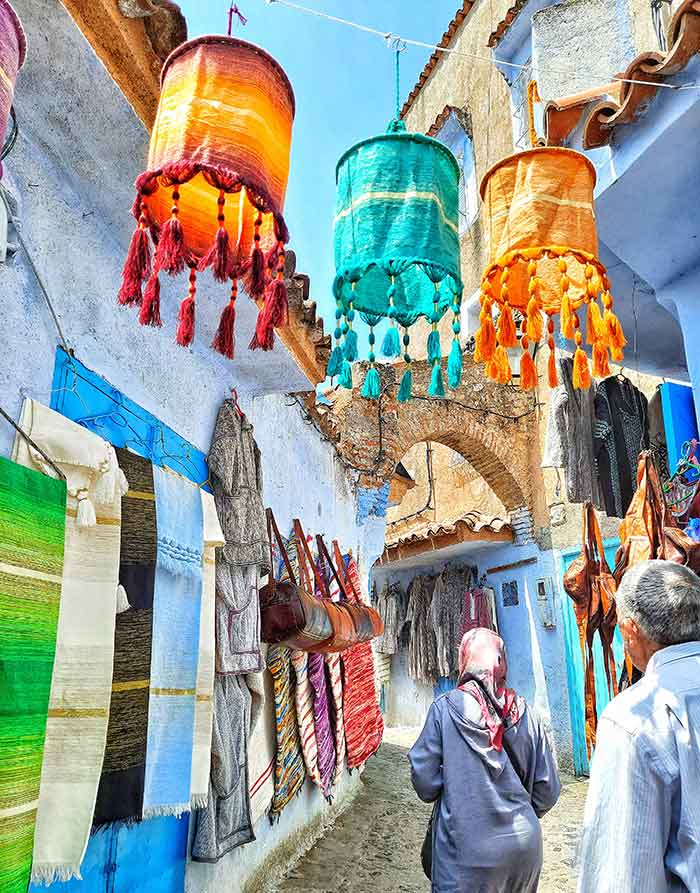 Two visitors stroll along the alleys of Chefchaouen streets, where colorful hanging lanterns, shawls and bags placed on the streets