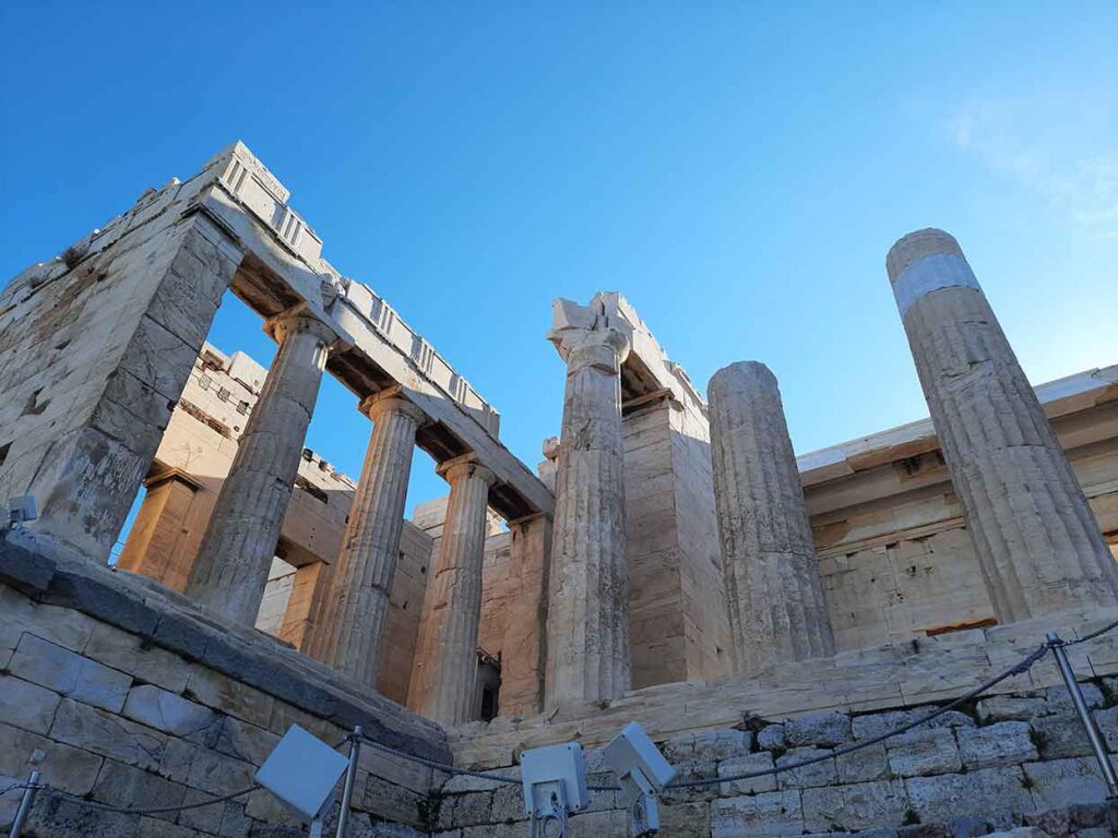 Entrance at the Acropolis features the temple with columns and cobblestone in an off-white color under a blue sky in the background