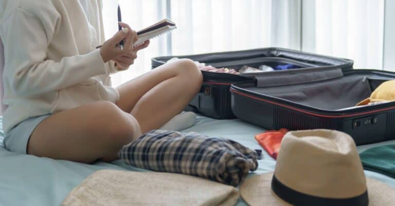 Woman packing a suitcase on a bed while checking a notebook for a last minute travel checklist