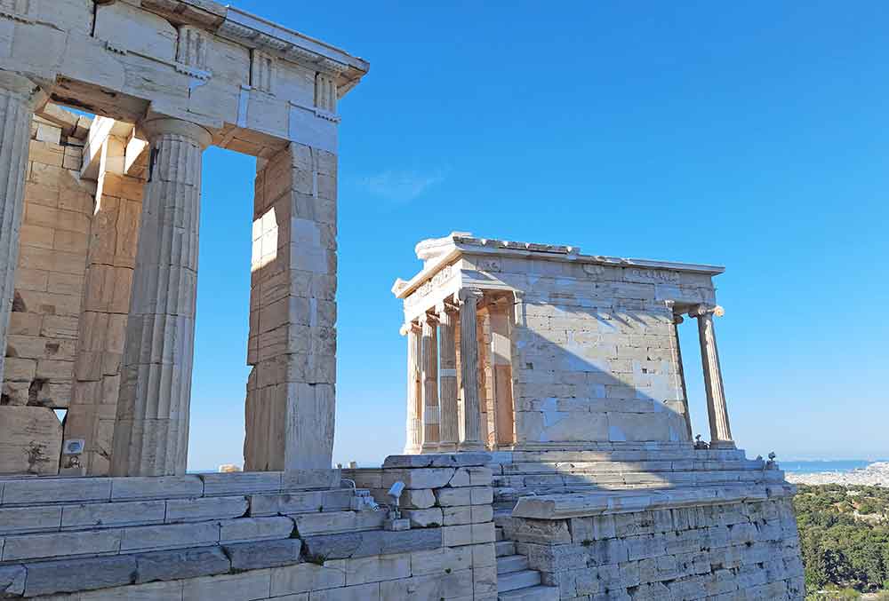 The Temple of Athena Nike features white-brown columns and bricks with a clear blue sky in the background