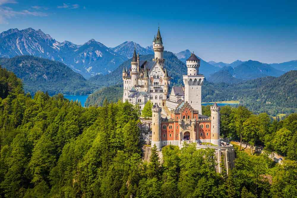 Neuschwanstein Castle in Germany features a stunning white and peach limestone facade, with towers crowned with pointed roofs, surrounded by lush greenery and offers panoramic views of mountains, forests, and lakes