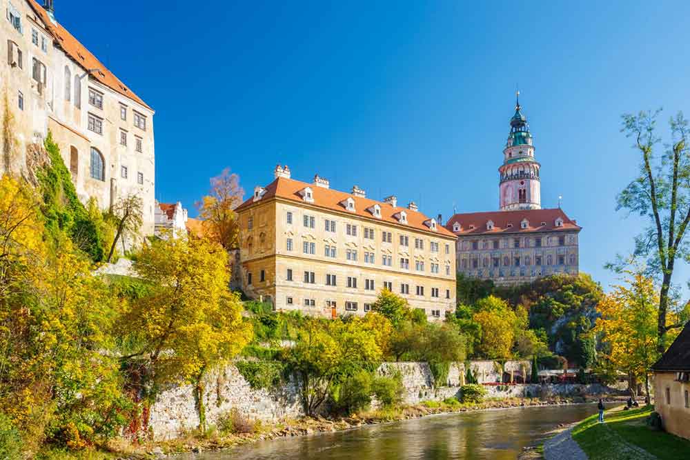 Cesky Krumlov Castle , Czech Republic features an orange roof and peach walls with multiple windows surrounded by trees and lake on the side 