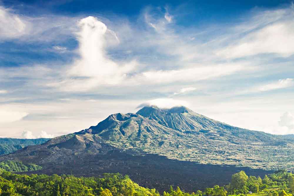 Mount Batur in Bali - a volcano with almost a symmetrical shape surround by trees in a white-blue sky