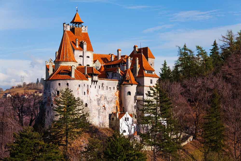 Bran Castle in Romania stands atop a rocky hill surrounded by trees - the castle showcases a red-orange roof and gray high walls against a blue sky background