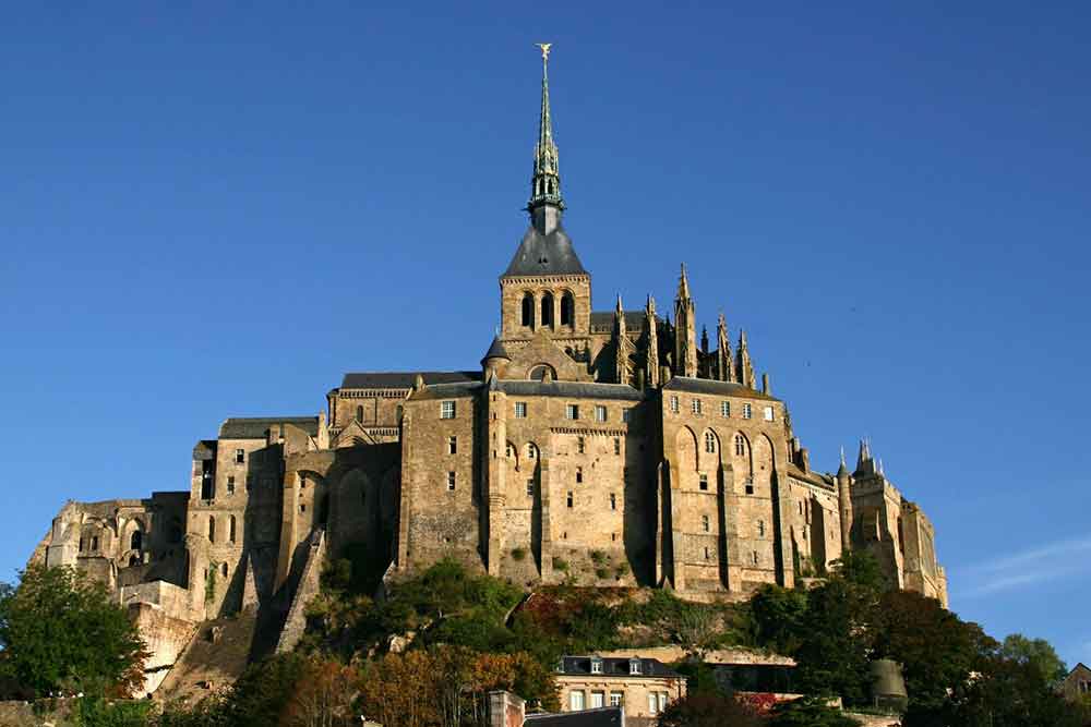 Mont Saint-Michel, France stands in a rocky hill with its towering silhouette and crowned by the abbey spire against the backdrop of the blue sky